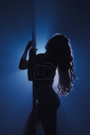 Foto de Silueta oscura de mujer flexible y plástica bailando sobre asta. Bailarina con pelo largo posando sobre pilón en estudio oscuro contra proyector brillante - Imagen libre de derechos