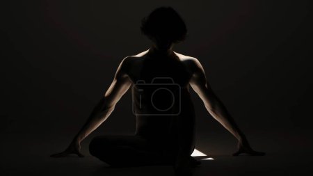 Foto de Body beauty and healthcare creative advertisement concept, Retrato de modelo masculino en estudio sobre fondo negro bajo el foco. Atractiva silueta de hombre posando en el suelo ante la cámara. - Imagen libre de derechos