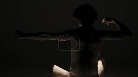 Photo for Body beauty and healthcare creative advertisement concept, Portrait of male model in studio on the black background under spotlight. Attractive man silhouette posing hands up. Back view - Royalty Free Image