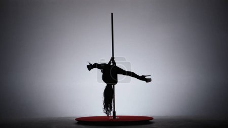Foto de Baile de poste. Una silueta de una chica interpretando elementos acrobáticos en un pilón en un estudio oscuro a la luz de focos sobre un fondo blanco - Imagen libre de derechos