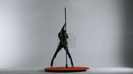 Téléchargez les photos : Pole dance. Une silhouette d'une fille exécutant des éléments acrobatiques sur un pylône dans un studio sombre à la lumière de projecteurs sur un fond blanc - en image libre de droit