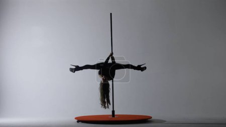 Téléchargez les photos : Pole dance. Une silhouette d'une fille exécutant des éléments acrobatiques sur un pylône dans un studio sombre à la lumière de projecteurs sur un fond blanc - en image libre de droit