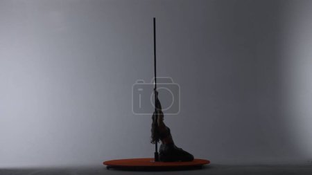 Photo for Pole dance. A silhouette of a girl performing acrobatic elements on a pylon in a dark studio in the light of spotlights on a white background - Royalty Free Image