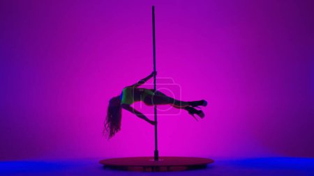 Photo for Young spectacular woman shows her flexibility and plasticity during pole dancing. Woman in yellow net suit dancing on pylon in studio on pink background with blue light - Royalty Free Image
