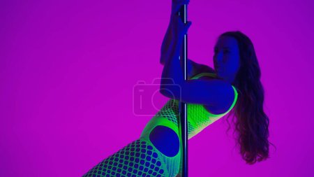 Foto de Joven mujer espectacular muestra su flexibilidad y plasticidad durante el baile de polo. Mujer en traje de red amarillo bailando sobre pilón en estudio sobre fondo rosa con luz azul - Imagen libre de derechos