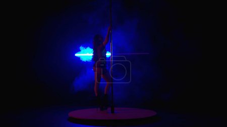 Foto de Baile de poste. Mujer joven bailando en un estudio oscuro sobre un fondo de luces de neón azul en bocanadas de humo. Mujer bailando striptease. Mujer sexy en zapatos de tacón alto - Imagen libre de derechos
