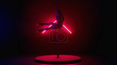 Téléchargez les photos : Silhouette d'une jeune femme exécutant des tours sur un poteau. Danseuse sur talons hauts démontrant des éléments de danse en pylône dans un studio sombre avec lumière rouge et fumée - en image libre de droit