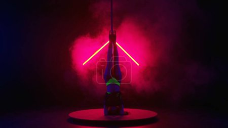 Photo for Silhouette of a young woman performing tricks on a pole. Dancer on high heels demonstrating elements of pylon dance in dark studio with red light and smoke - Royalty Free Image