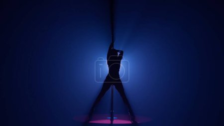 Foto de Silueta oscura de una mujer flexible y plástica bailando sobre un poste. Mujer bailando sobre un pilón en un estudio oscuro contra un reflector brillante - Imagen libre de derechos