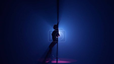Foto de Silueta oscura de una mujer flexible y plástica bailando sobre un poste. Mujer bailando sobre un pilón en un estudio oscuro contra un reflector brillante - Imagen libre de derechos