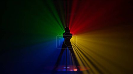 Photo for Dark silhouette of flexible and plastic woman dancing on a pole. A woman dancing on a pylon in a dark studio, illuminated by multicolored beams of a spotlight - Royalty Free Image