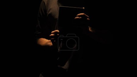 Photo for Historical and vintage objects creative advertisement concept. Studio shot of old retro hardcover book on dark background in warm light. Man holding closed old vintage book in hands in darkness. - Royalty Free Image