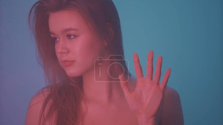Photo for Beauty and cosmetology creative advertisement concept. Portrait of female in neon light behind the glass window in steam mist. Girl with makeup and natural hairstyle holds hand on glass. - Royalty Free Image