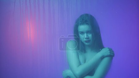 Téléchargez les photos : Beauté et cosmétologie concept publicitaire créatif. Portrait de femme au néon derrière la vitre en vapeur et gouttes d'eau. Fille avec maquillage et coiffure naturelle posant elle-même étreinte. - en image libre de droit