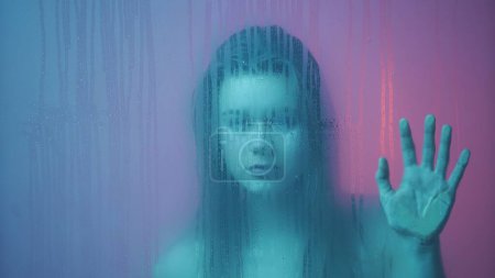 Téléchargez les photos : Concept de publicité créative beauté. Portrait de femme au néon derrière la vitre en vapeur et gouttes d'eau. Fille avec maquillage et coiffure, les mains sur le verre regarde dans la caméra. - en image libre de droit