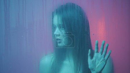 Photo for Beauty and cosmetology creative advertisement concept. Portrait of female in neon light behind the glass window in steam and water drops. Girl with makeup and hairstyle, hands on glass looks around. - Royalty Free Image