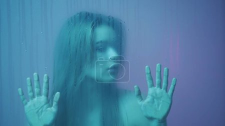 Photo for Beauty and cosmetology creative advertisement concept. Portrait of female in neon light behind the glass window in steam and water drops. Girl with makeup and hairstyle, hands on glass looks around. - Royalty Free Image