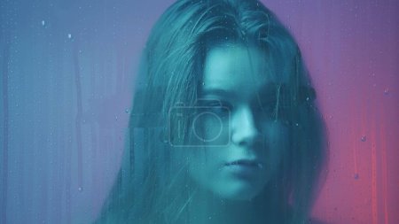 Photo for Beauty and cosmetology creative advertisement concept. Portrait of female in neon light behind the glass window in steam and water drops. Girl with makeup looks around at camera through wet glass. - Royalty Free Image