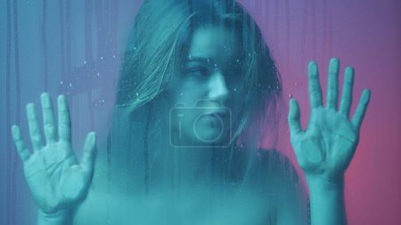 Téléchargez les photos : Beauté et cosmétologie concept publicitaire créatif. Portrait de femme au néon derrière la vitre en vapeur et gouttes d'eau. Attrayant fille tenant la main sur le verre et regardant à travers. - en image libre de droit