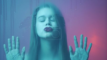 Photo for Beauty and cosmetology creative advertisement concept. Portrait of female in neon light behind the glass window in steam and water drops. Girl with makeup and natural hairstyle kissed the wet glass. - Royalty Free Image