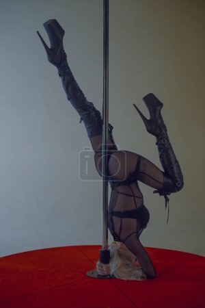 Photo for Young blonde woman in black tight jumpsuit and high-heeled boots performs headstand near pylon. Dancer performs on the podium of the pylon on the white background of the studio - Royalty Free Image