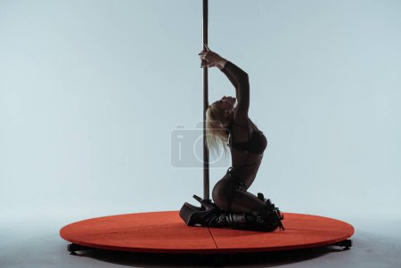 Photo for Young blonde woman in black tight jumpsuit and high heeled boots poses sitting on catwalk near pylon. Dancer performs on white background of studio, exotic dance - Royalty Free Image