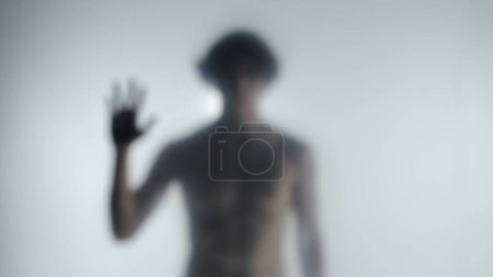 Téléchargez les photos : Silhouette floue d'un homme avec un torse nu derrière un rideau ou un verre givré. L'homme touche le rideau transparent avec sa main et regarde dans la caméra. Concept de fantôme - en image libre de droit