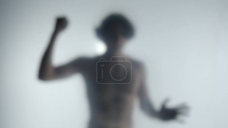 Foto de Una silueta borrosa de un hombre con un torso desnudo detrás de una cortina esmerilada o vidrio. Un hombre golpeando su puño, tratando de romper una barrera invisible, inclinando su cabeza en la desesperanza. Concepto fantasma - Imagen libre de derechos