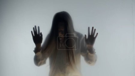Téléchargez les photos : Silhouette de femme paniquée dans le brouillard derrière un verre givré ou un rideau. La femme se cogne la tête contre une barrière transparente et invisible. Concept de l'au-delà et de l'au-delà, fantômes et esprits - en image libre de droit