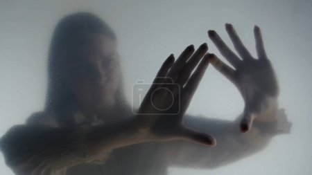 Téléchargez les photos : Silhouette de femme fantôme dans le brouillard derrière rideau givré ou verre fermer. Une femme touche le verre. Concept d'au-delà, autre monde, fantômes - en image libre de droit