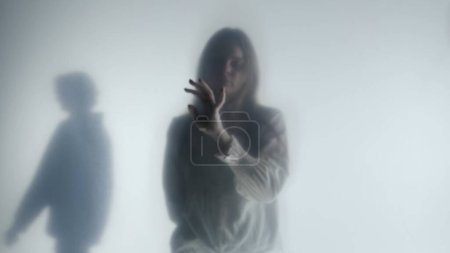 Silhouette of a man and a woman behind a frosted curtain or glass. The woman is pensively touching the curtain with her hands, the man is visible in the background
