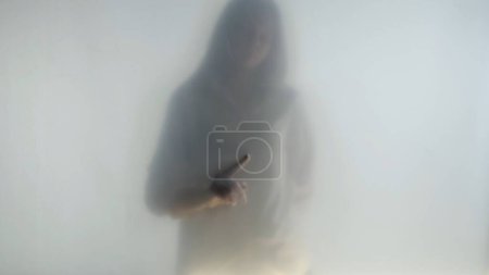 Téléchargez les photos : Silhouette de femme fantôme dans le brouillard derrière rideau givré ou verre fermer. La femme dessine des motifs invisibles sur le verre avec son index. Le concept de l'au-delà, l'au-delà, les fantômes - en image libre de droit