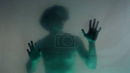 Téléchargez les photos : Une silhouette floue d'un homme au torse nu derrière un rideau mat, au néon vert. L'homme touche la barrière transparente, essayant de se libérer. Le concept des fantômes et des esprits - en image libre de droit