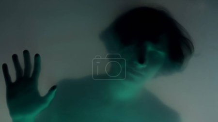 Foto de Una silueta borrosa de un hombre con un torso desnudo detrás de una cortina mate, con luz de neón verde de cerca. El hombre está tocando la barrera transparente, intentando liberarse. El concepto de fantasmas y - Imagen libre de derechos
