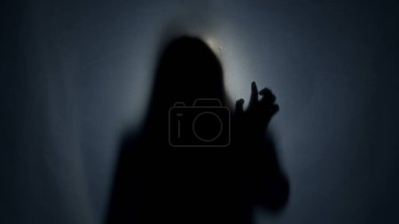 Foto de Una silueta oscura de una mujer detrás de una cortina de estera, iluminada por un haz de luz. La mujer toca la cortina. La otra vida, el otro mundo - Imagen libre de derechos
