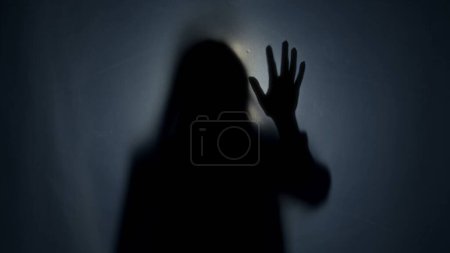 Foto de Una silueta oscura de una mujer detrás de una cortina de estera, iluminada por un haz de luz. La mujer toca la cortina. La otra vida, el otro mundo - Imagen libre de derechos