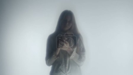 Téléchargez les photos : Silhouette d'une fille en vêtements blancs derrière un rideau ou un verre givré. Une femme triste se tient la tête baissée. Concept mystique - en image libre de droit