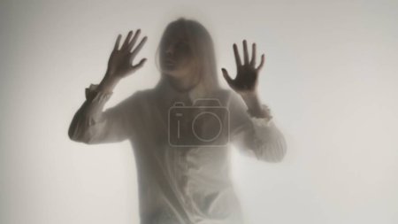 Foto de Una silueta femenina melancólica detrás de una cortina o vidrio esmerilado transparente. Una mujer toca la superficie de la cortina con sus palmas - Imagen libre de derechos
