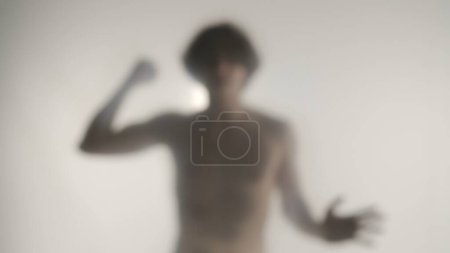 Téléchargez les photos : Une silhouette floue d'un homme avec un torse nu derrière un rideau ou un verre givré. Un homme frappant son poing, essayant de briser une barrière invisible, inclinant la tête dans le désespoir. Concept de fantôme - en image libre de droit