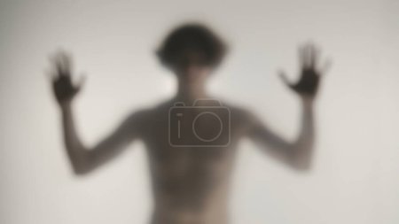 Téléchargez les photos : Silhouette floue d'un homme avec un torse nu derrière un rideau ou un verre givré. L'homme touche le rideau transparent avec deux paumes et regarde dans la caméra - en image libre de droit
