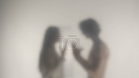 Téléchargez les photos : Silhouette d'un couple qui se dispute derrière un verre ou un rideau givré. Une femme et un homme se querellent et gesticulent émotionnellement avec leurs mains - en image libre de droit