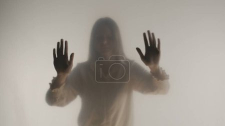 Téléchargez les photos : Une silhouette féminine mélancolique derrière un rideau ou un verre dépoli transparent. Une femme touche la surface du rideau avec ses paumes - en image libre de droit