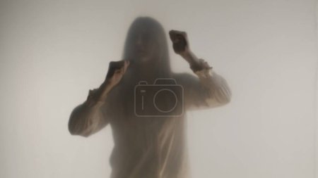Téléchargez les photos : Silhouette de femme paniquée dans le brouillard derrière un verre givré ou un rideau. La femme frappe le verre avec ses poings pour tenter de briser la barrière - en image libre de droit