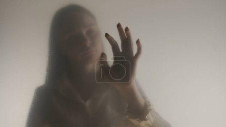Téléchargez les photos : Silhouette de femme fantôme dans le brouillard derrière rideau givré ou verre fermer. Une femme touche le verre. Concept d'au-delà, autre monde, fantômes - en image libre de droit