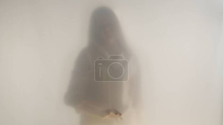 Téléchargez les photos : Silhouette floue d'une femme vêtue de blanc derrière un rideau ou un verre givré - en image libre de droit