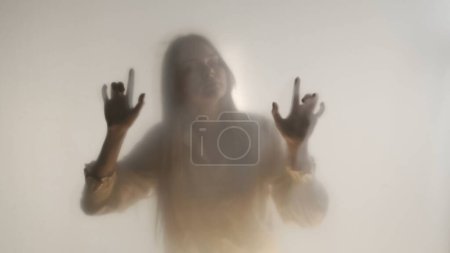 Téléchargez les photos : Silhouette féminine mélancolique derrière un rideau givré transparent ou un verre fermé. Une femme guide sa main sur la surface du rideau - en image libre de droit