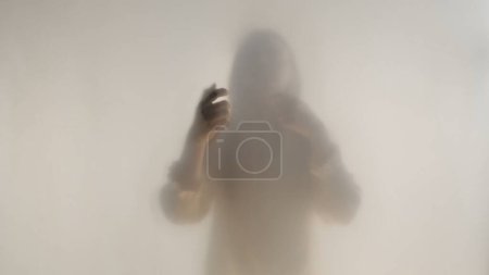 Téléchargez les photos : Silhouette féminine mélancolique derrière un rideau givré transparent ou un verre fermé. Une femme guide sa main sur la surface du rideau - en image libre de droit