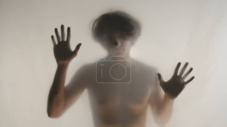 Téléchargez les photos : Silhouette floue d'un homme avec un torse nu derrière un rideau ou un verre givré. L'homme touche le rideau transparent avec deux paumes et regarde dans la caméra - en image libre de droit