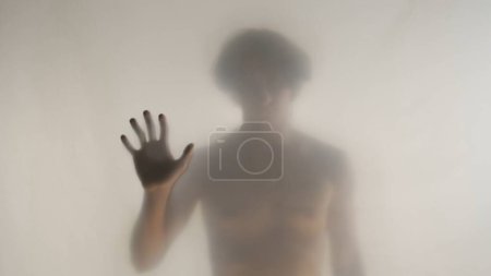 Foto de Silueta borrosa de un hombre con un torso desnudo detrás de una cortina esmerilada o vidrio. El hombre toca la cortina transparente con dos palmas y mira a la cámara - Imagen libre de derechos