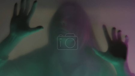 Foto de Silueta borrosa de una mujer detrás de una cortina de estera, en luz de neón rosa y verde, de cerca. Una mujer se está raspando las uñas en la barrera, tratando de romperla. El concepto de fantasmas y espíritus - Imagen libre de derechos
